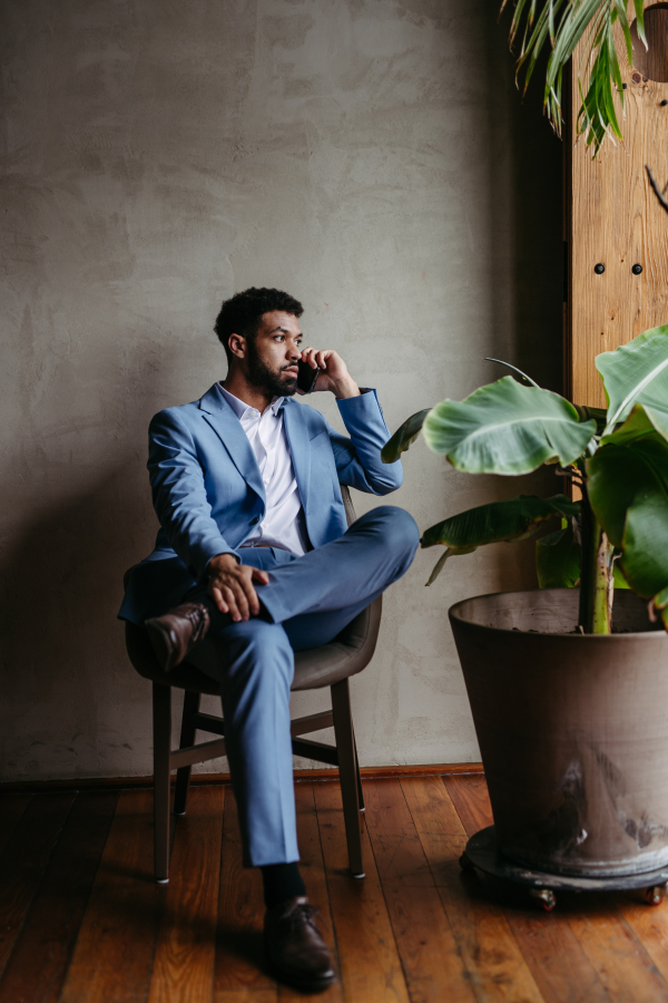 Portrait of a young businessman sitting near window in modern office full of plants. Male corporate leader, executive, manager making phone call.