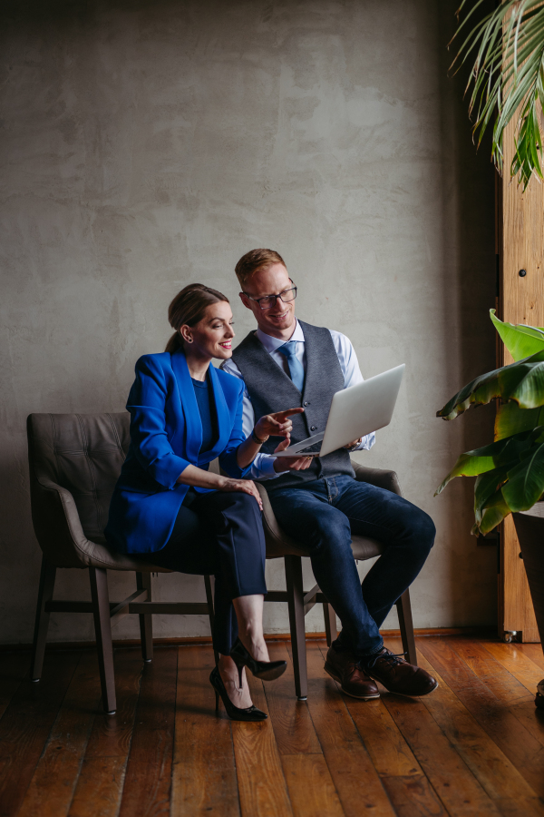 Two colleagues sitting near window and working on notebook in modern office full of plants. Male and female manager teamworking.