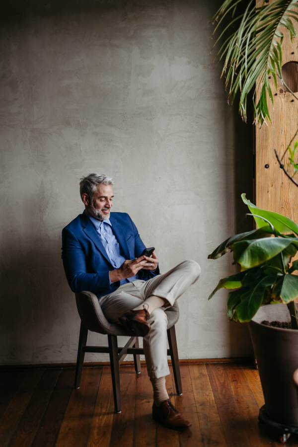 Portrait of a businessman sitting near window in modern office full of plants. Male corporate leader, executive, manager using smart phone..