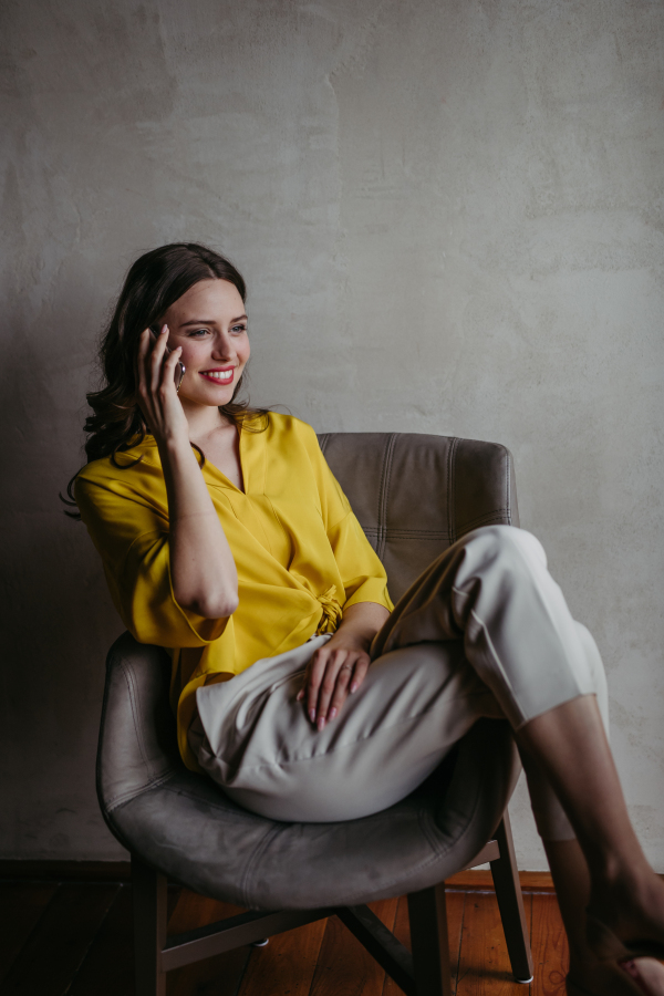 Portrait of a young businesswoman sitting near window in modern office full of plants. Female corporate leader, executive, manager making phone call.