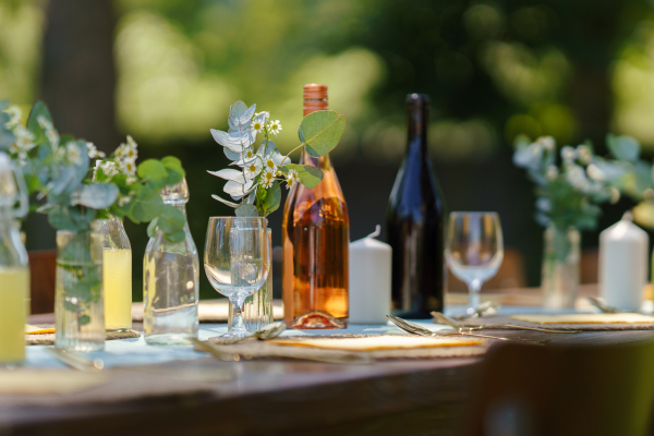 Close up shot of a set table at a summer garden party. Table setting with glasses, lemonade, delicate floral decoration, and bottles of summer wine.