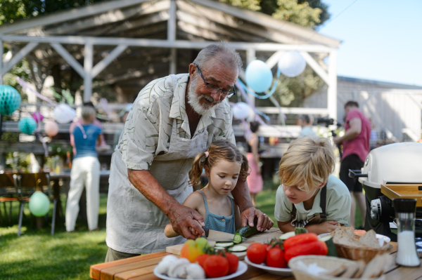 Grandfather teaching kids to cut vegetables for grilling. Grilling together at garden BBQ. Grandchildren with grandfatehr at summer family garden party.