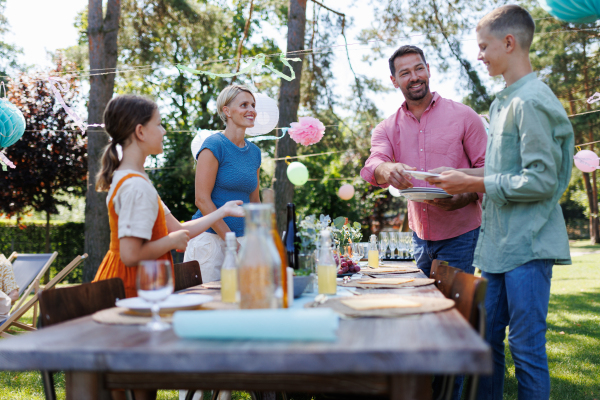 Family setting table for summer garden party. Bringing plates, food, and drinks.