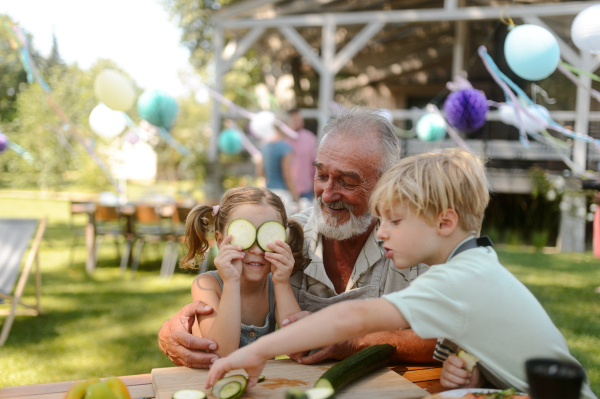 Grandfather and grandchildren having fun at a garden barbecue. Family gathering at summer garden party. Granddauhter putting cucumber slices on her eyes.