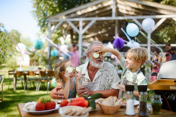 Grandfather and grandchildren having fun at a garden barbecue. Family gathering at summer garden party. Grandson putting cucumber slices on his grandpa's eyes.