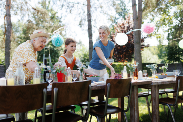 Grandmother, mother and daughter setting table for summer garden party. Three generations of women in family, bringing plates, food and drinks at party table. Getting ready for family gathering.