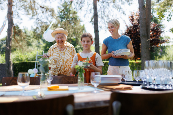 Grandmother, mother and daughter setting table for summer garden party. Bringing plates, food, and drinks.