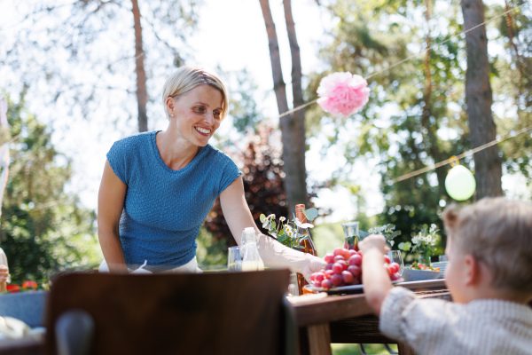 Smiling mother giving little boy grape berry. Family garden party with a set table.