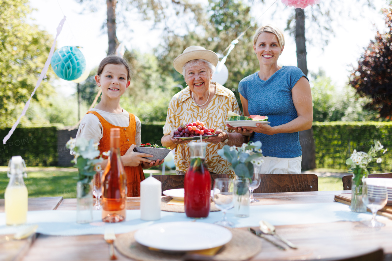 Grandmother, mother and daughter setting table for summer garden party. Three generations of women in family, bringing plates, food, and drinks at table.