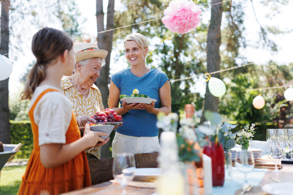 Girl helping grandmother and mother to set table for summer garden party. Bringing plates, food, and drinks. Tree generations of woman in family.