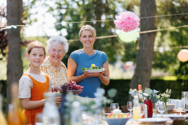 Grandmother, mother and daughter setting table for summer garden party. Three generations of women in family, bringing plates, food, and drinks at table.