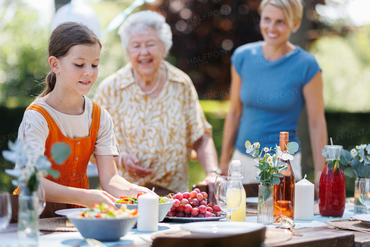 Grandmother, mother and daughter setting table for summer garden party. Bringing plates, food, and drinks.