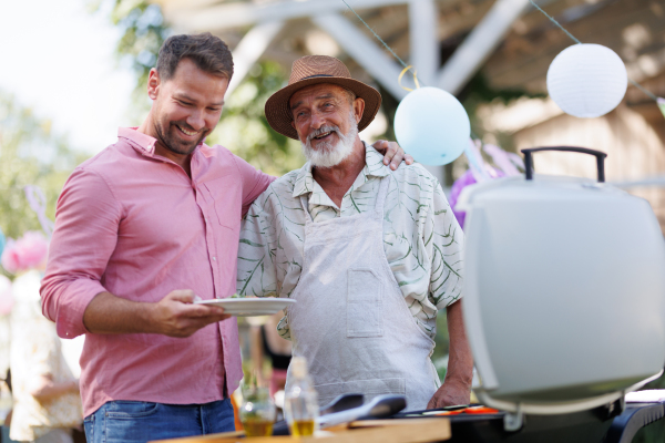 Senior father and mature son grilling together at a garden bbq party. Man looking at full plate with grilled food.