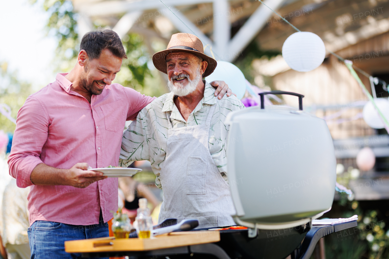 Senior father and mature son grilling together at a garden bbq party. Man looking at full plate with grilled food.