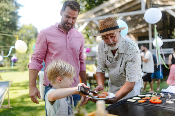 Father, grandfather and son grilling together at a garden bbq party. Three generations of men at summer family garden party.