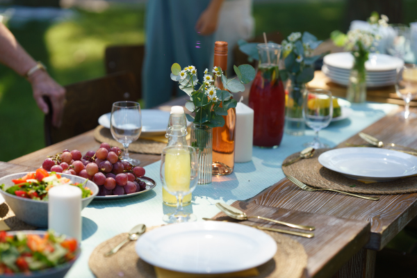 Close up shot of a set table at summer garden party, grilled food. Table setting with glasses, lemonade, delicate floral and paper decoration, and bottles of summer wine.