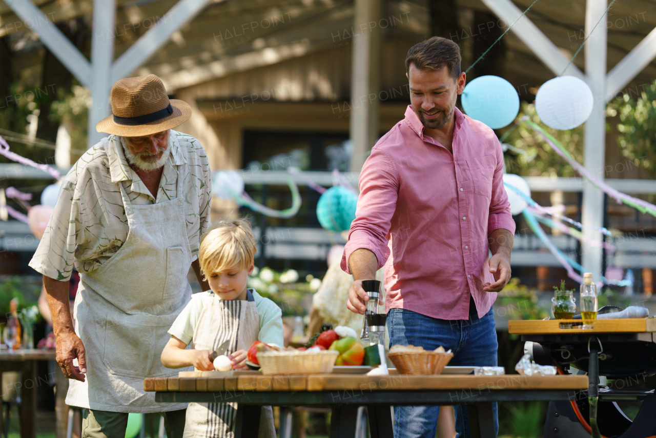 Young boy cutting vegetables for grilling. Father, grandfather and son grilling together at a garden bbq party. Three generations of men at summer family garden party.