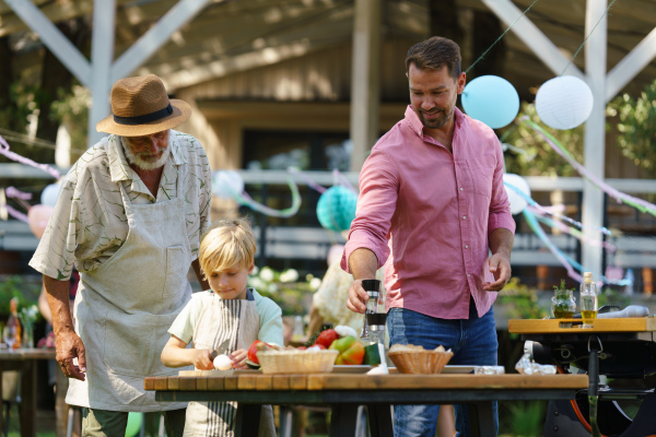 Young boy cutting vegetables for grilling. Father, grandfather and son grilling together at a garden bbq party. Three generations of men at summer family garden party.