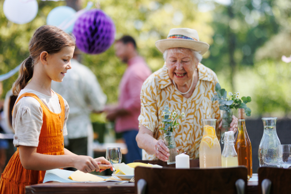 Girl helping grandmother to set table for summer garden party. Bringing plates, food, and drinks.