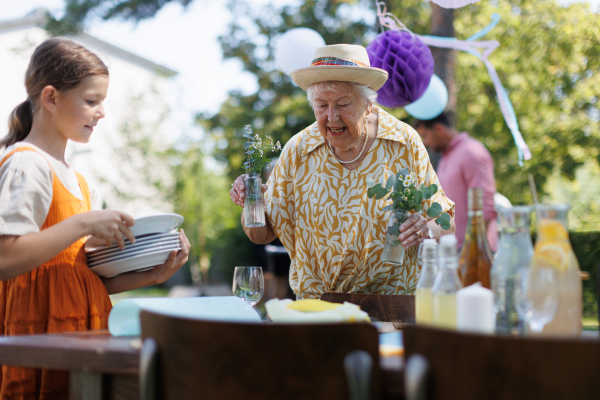 Grandmother and granddaughter setting table for summer garden party. Bringing plates and flower decoration.