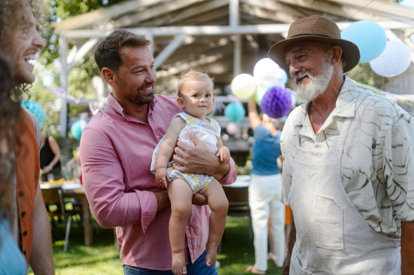 Father holding little baby in arms, talking with babys grandfather and family. Family summer garden party.