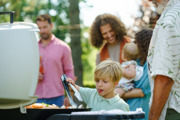 Young boy turning grilled food with tongs. Grandfather and grandson are grilling together at a garden bbq party.