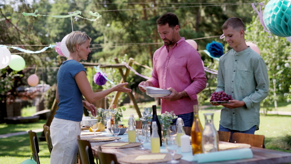 Family setting table for summer garden party. Bringing plates, food, and drinks.