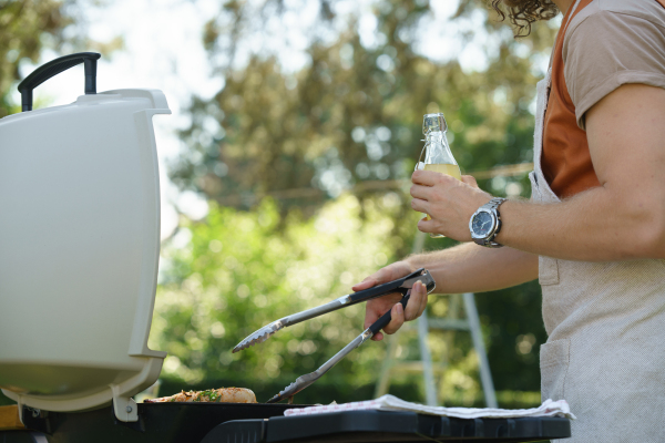 Man grilling on outdoor grill, holding tongs and drinkig lemonade. Outdoor grill or BBQ party in the garden.