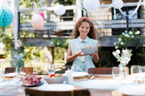 Girl helping to set table for summer garden party. Bringing plates, food, and drinks.