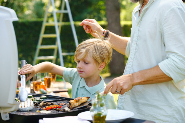 Young boy turning grilled cheese with tongs. Father and son grilling together at a garden bbq party.