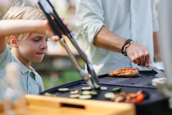 Young boy turning grilled vegetables with tongs. Grandfather and grandson are grilling together at a garden bbq party.