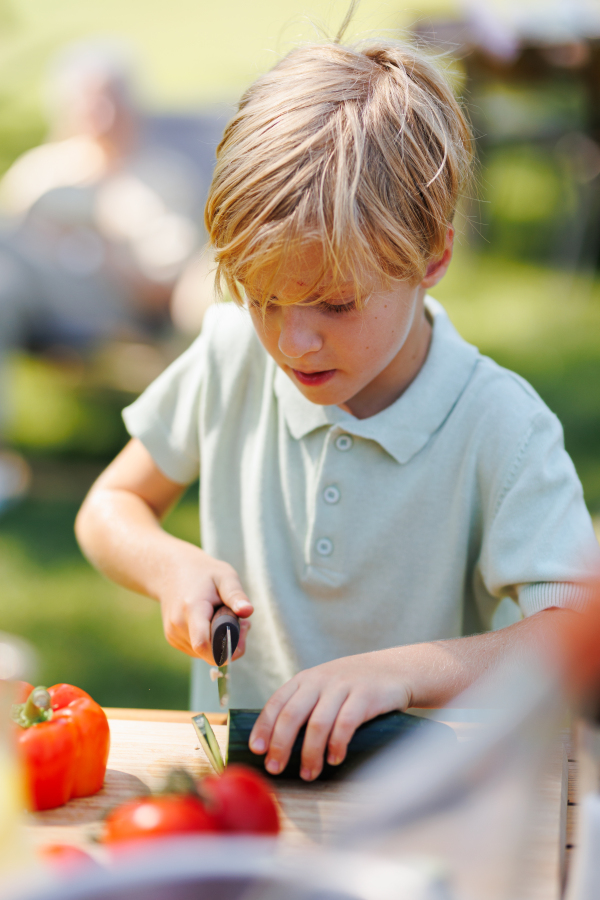 Young boy cutting vegetables for grilling. Father, grandfather and son grilling together at a garden bbq party. Three generations of men at summer family garden party.