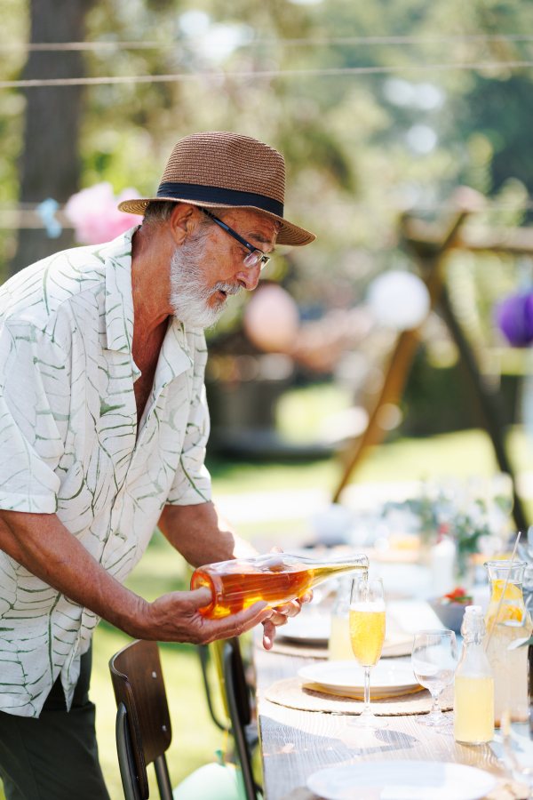 Elderly handsome man preparing refreshments for a summer garden party, pouring wine in glass.