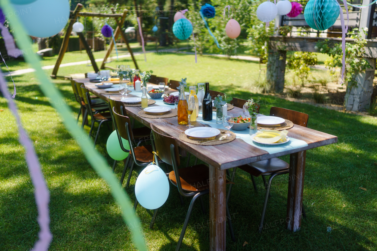 Close up shot of a set table at a summer garden party. Table setting with glasses, fruit lemonade, fresh fruits and salads and delicate floral decoration. Colorful paper decorations for party.