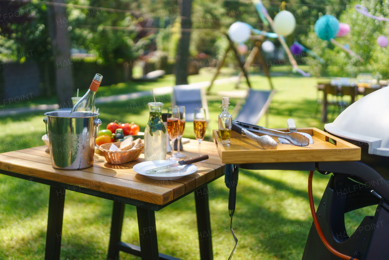 Close up shot of a grill and table at a summer garden party. Side table with wine glasses, lemonade, fresh vegetables aand bread.