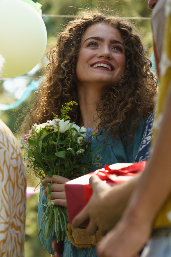 Portrait of surprised birthday girl at garden birthday party. Friends holding birthday gifts, flowers.