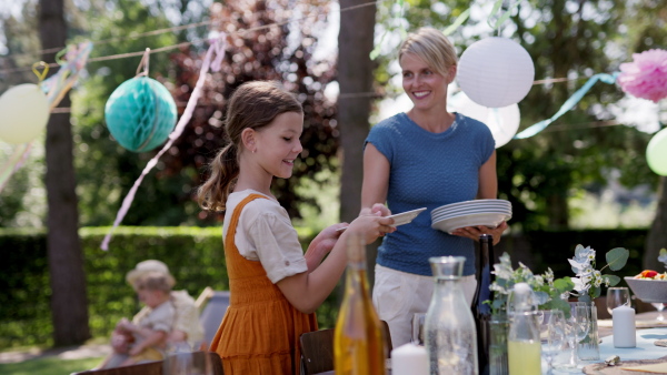 Mother and daughter setting table for summer garden party. Three generations of women in family, bringing plates, food, and drinks at table.