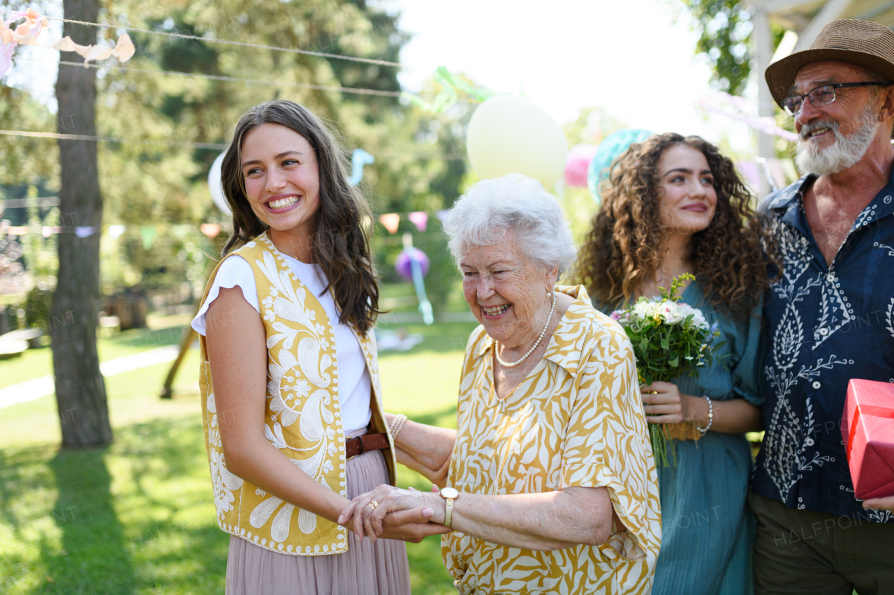 Beautiful senior birthday woman receiving gift from granddaughter. Two mature sisters with grandparents at birthday party. Multigenerational family.