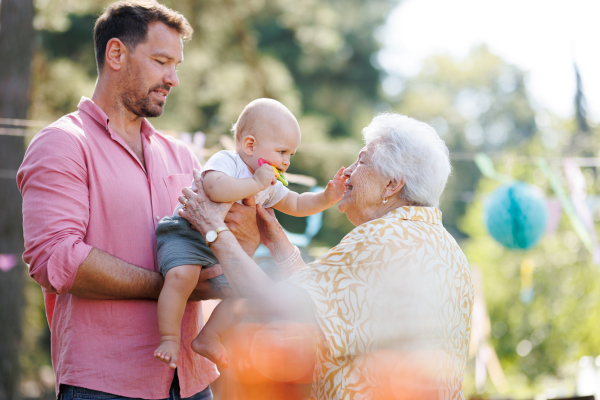 Great-grandmother holding little baby in her arms, father assisting. Family summer garden party.