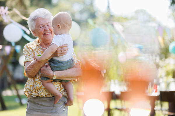 Portrait of great-grandmother holding a tiny baby in her arms. Family summer garden party.