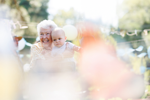 Portrait of great-grandmother holding a tiny baby in her arms. Family summer garden party.