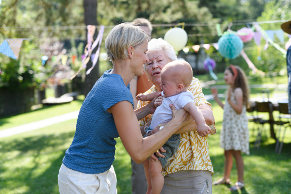 Great-grandmother holding crying little baby in her arms. Mother soothing fussy baby. Family summer garden party.