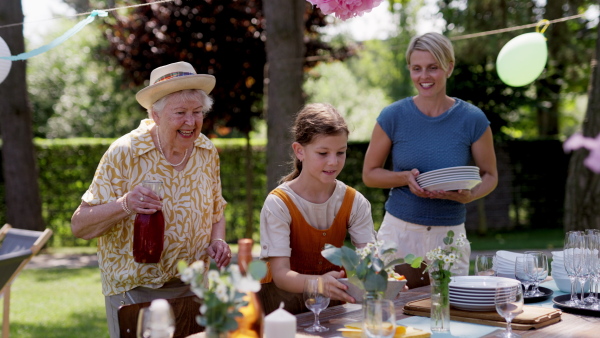 Grandmother, mother and daughter setting table for summer garden party. Three generations of women in family, bringing plates, food, and drinks at table.