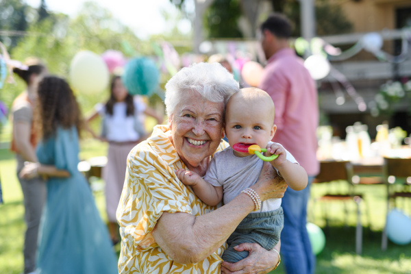 Portrait of great-grandmother holding a tiny baby in her arms. Family summer garden party.