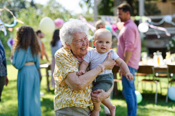 Portrait of great-grandmother holding a tiny baby in her arms. Family summer garden party.