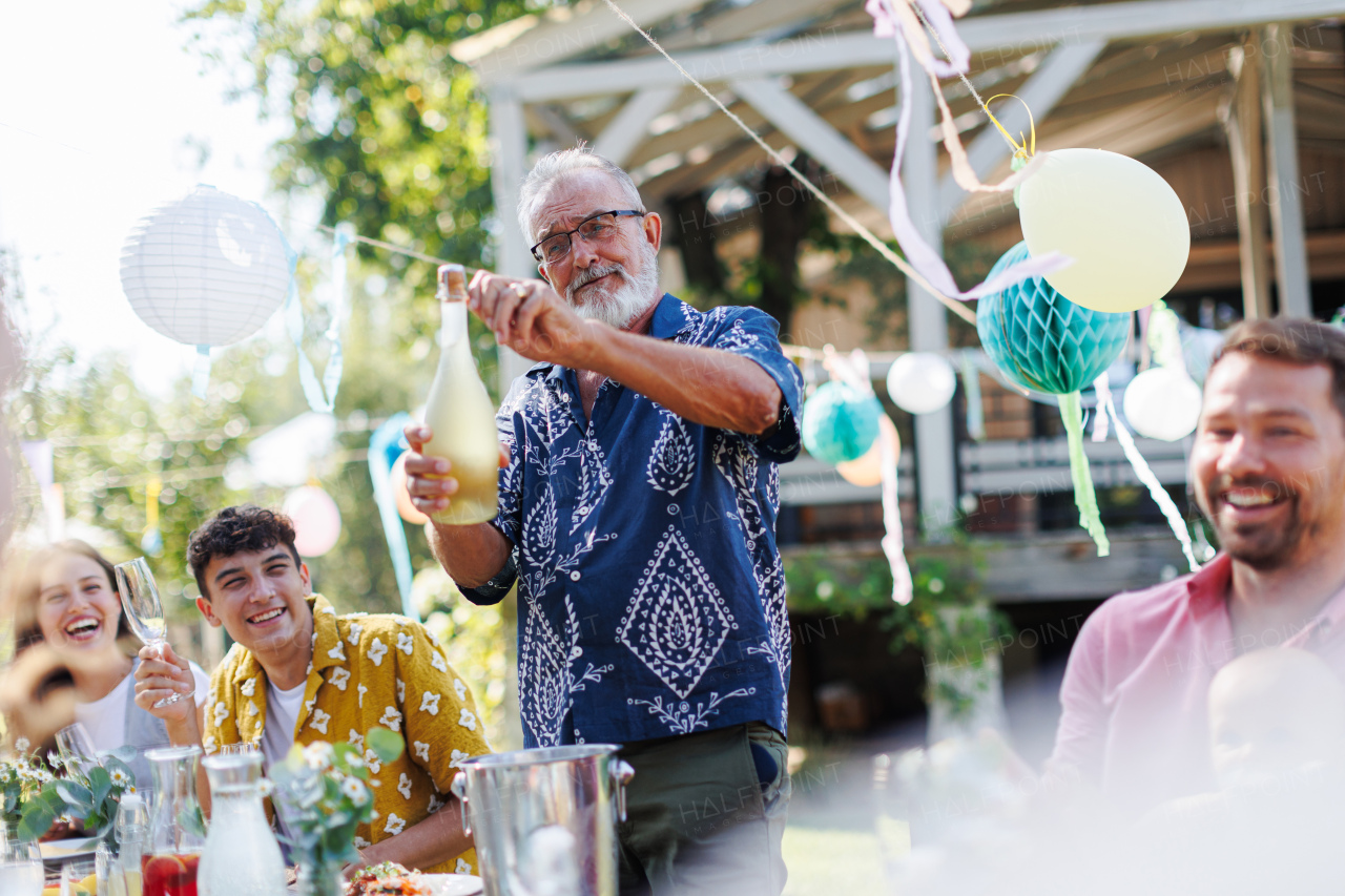 Grandfather opening a bottle of champagne, pouring a champagne into glasses. Senior man making a celebratory toast at an outdoor summer garden party.