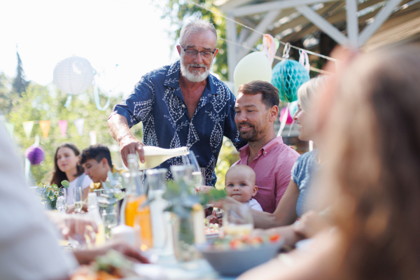 Grandfather opening a bottle of champagne, pouring wine into glasses. Senior man making a celebratory toast at an outdoor summer garden party.