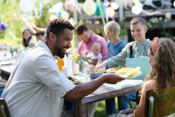 Man offering plate with food to little girl sitting at table. Eating grilled food outdoors. Family garden party, BBQ.