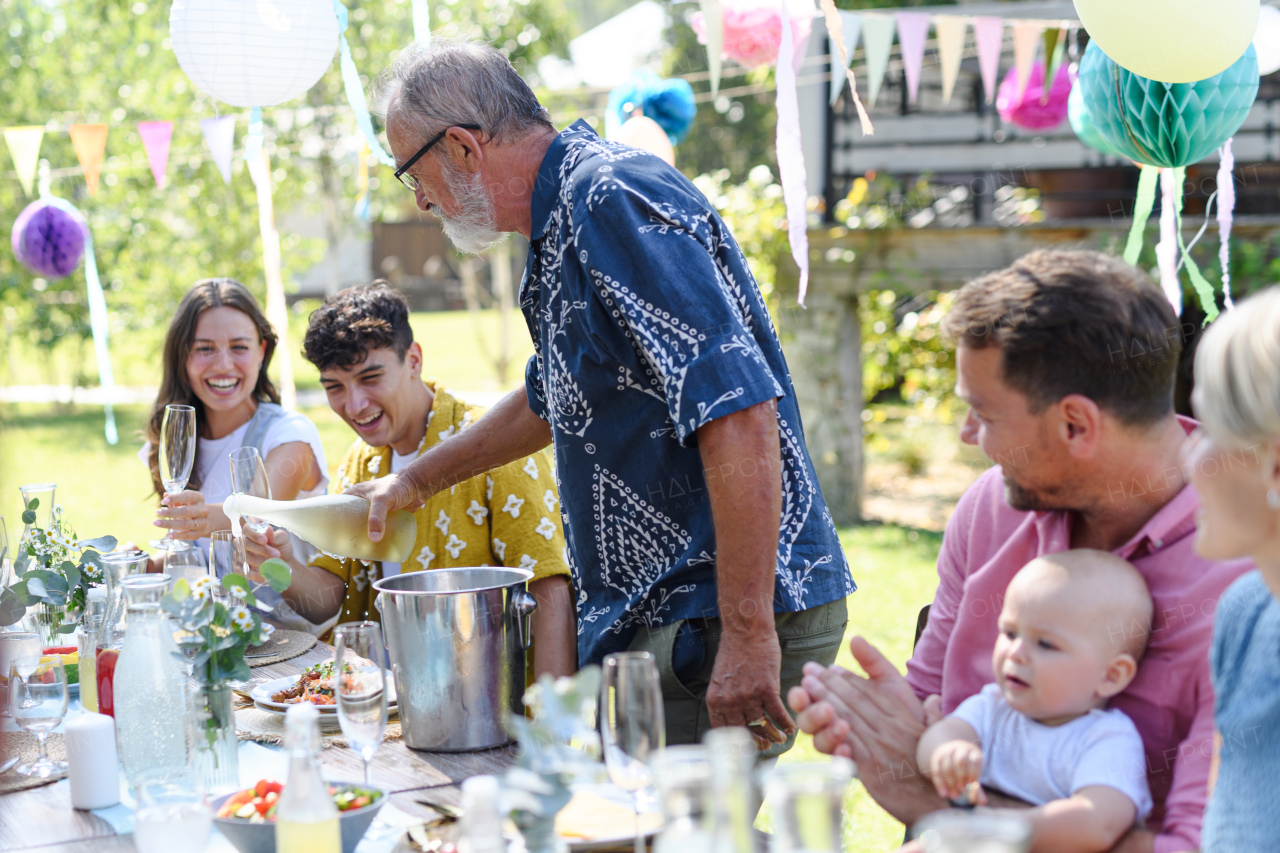 Grandfather opening a bottle of champagne, pouring a champagne into glasses. Senior man making a celebratory toast at an outdoor summer garden party.