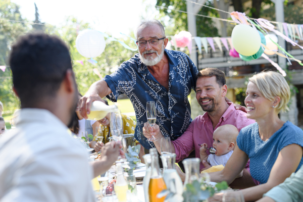 Grandfather opening a bottle of champagne, pouring a champagne into glasses. Senior man making a celebratory toast at an outdoor summer garden party.